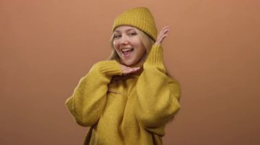 Sarı kazaklı ve bereli bir kadın turuncu arka planda gülümsüyor, elleri çenesinin altında neşeli bir yüz ifadesi sergiliyor..