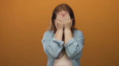 Kırmızı saçlı, mavi gömlekli, yüzü turuncu duvarlı, elleri stüdyoda olan bir kadın..