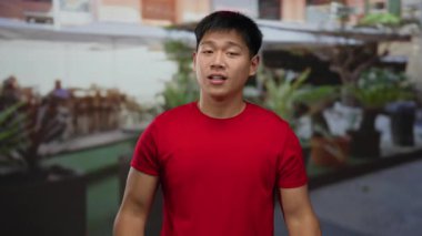 Kırmızı tişörtlü genç bir adam bir restoranın terasında hareketli bir kahve dükkanında canlı bir sohbet sunuyor..