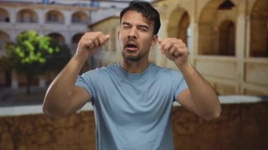 Genç İspanyol bir adam eski bir üniversite avlusunun önünde neşeli bir ağlama hareketi yapıyor, mizahi bir açık hava anını yakalıyor..