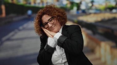 Orta yaşlı, esmer, kıvırcık saçlı ve gözlüklü bir kadın güneşli bir şehir sokağında siyah takım elbise ve beyaz gömlek giyip, yorgunluğunu ifade ederek uyku jesti yapıyor..