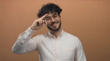 Beyaz gömlekli, sakallı, gözlüklü, turuncu arka planda poz veren, gülümseyen ve parmaklarını kafasında gezdiren genç bir İspanyol. Olumlu ve dostane bir hava yaratıyor..