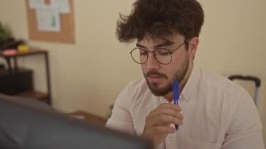 Ofisteki genç adam elinde kalemle bilgisayar işine odaklanmış, etrafı ofis dekoruyla çevrili, profesyonel ve düşünceli bir atmosfer sunuyor..