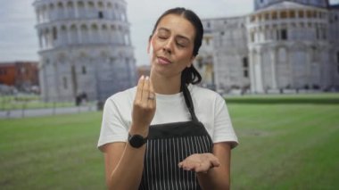 Çizgili önlüklü kadın çimdiklenmiş parmaklarını binanın önünde açık avuçla hareket ettiriyor. Düşünceli bir yansıma..