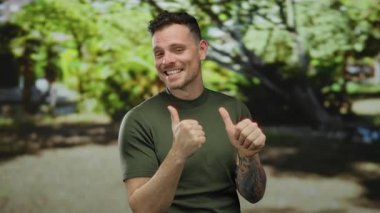 Parkta baş parmağıyla gülümseyen genç bir adam açık havada neşeli ve kendinden emin bir yüz ifadesi sergiliyor yemyeşil ağaçlarla çevrili aydınlık bir günde..