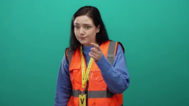 Gönüllü yelek giymiş esmer bir kadın yeşil bir zemine karşı ezik jestler yapıyor..