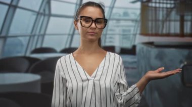 Gözlüklü ve çizgili bluz giyen genç bir kadın, restoranın iç kısımlarında gülümseyerek ve jest yaparak avuç içini işaret ediyor;.