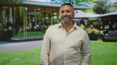 Sakallı bir adam gülümsüyor ve cam bir binanın önünde duruyor bej bir keten gömlek giyiyor, rahat duruş; sıcak yaklaşılabilir memnuniyet.