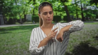 At kuyruklu kadın orman parkında çıplak bileğini işaret ediyor, saati gösteriyor ve sarı tırnaklı jestleri kontrol ediyor;.