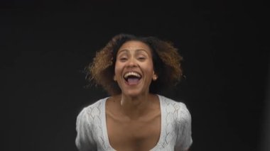 Siyahi stüdyoda yumruklarını sıkmış ve kollarını kaldırmış beyaz örgü bluz giymiş ve atlarken geniş bir gülümseme takınan heyecanlı zafer güçlendirici kadın..