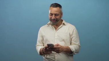 Orta yaşlı, gri sakallı, akıllı telefonu iki eliyle tutan ve mavi stüdyoda gülümseyen bir adam..