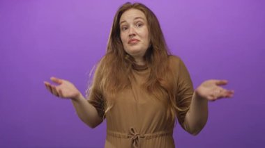 Stüdyoda mora karşı omuz silkme hareketleriyle elini kaldıran kadın kahverengi elbise giyiyor ve belirsizlik ifadesi sergiliyor..
