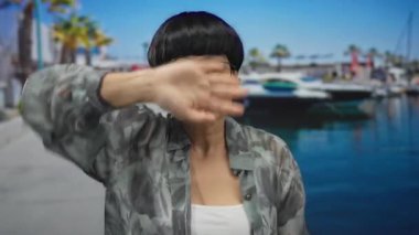 Elleri deniz kıyısında, açık mavi gökyüzünün altında tekneleri olan gözlüklü ve kamuflajlı bir ceket giyen bir kadın, açık havada anlamlı bir an sergiliyor..