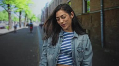 Shaka el hareketi yapan ve kot ceket ve çizgili ekin bluzu giyen, sokakta başparmağına işaret eden bir kadın..