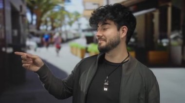 Sakallı genç bir adam, kalabalık bir caddede, sakin bir şehir ortamında güvenle gülümserken bir telefon konuşması yapıyor..