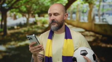Sakallı kel adam akıllı telefon ve futbol topu tutuyor, sarı ve mor atkı takıyor, güneşli bir parkta duruyor..