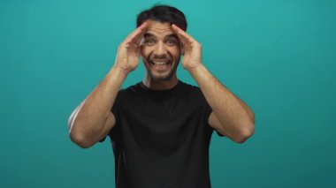 Siyah gömlekli genç adam, elleri turkuaz arka planda poz veriyor, stresi ve tefekkür etmeyi ifade ediyor..