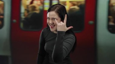 Kaya jesti yapan bir kadın koyu renk saçlı ve cesur ifadeli yeraltı jestleri yapıyor, tren arka planda bulanık, şehir sahnesinde canlı enerji sergiliyor..