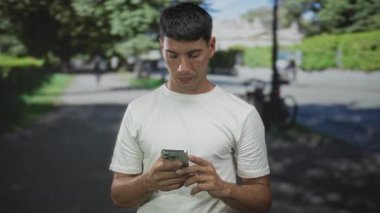 Beyaz tişörtlü adam, güneşli yeşil parkta el kamerasıyla mesajlaşan akıllı telefonu tutuyor..