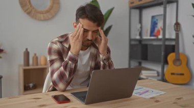 Genç adam, modern bir ofiste dizüstü bilgisayarı ve telefonu olan, bitkiler ve dekorlarla çevrili, tipik bir çağdaş çalışma ortamını gösteren stresli bir genç..