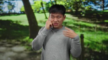 Genç adam güneşli bir parkta telefonla konuşuyor, duygularını canlı bir şekilde ifade ediyor, yemyeşil ağaçlarla çevrili.