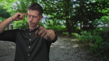 Kısa saçlı, bileklikli, parmakları şakaktan şakağa gezen ve ormanın kamerasını işaret eden beyaz bir adam..