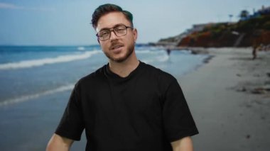 Kumsalda sakallı genç bir adam, açık gökyüzünün altında deniz kenarında el kol hareketi yapıyor. Açık hava ortamında karışık duyguları yansıtıyor..