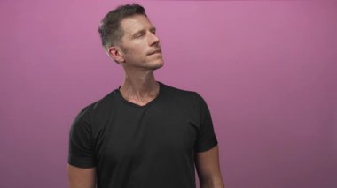Kafası öne eğilmiş, boynu açık gülümseyen, pembe stüdyoda siyah tişört giyen, kendine güveni tam genç beyaz bir adam..