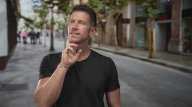 Adam, genç beyaz, şehir caddesinde çene üzerinde düşünme pozisyonu, siyah t-shirt ve bilezik giyiyor; tefekkür.