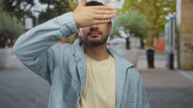 Açık mavi ceketli ve bej gömlekli bir adam sokak köşesinde yüzünü buruştururken gözlerini kapar; utangaçlık.