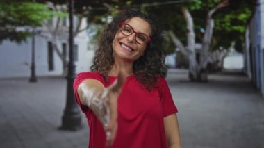 Kırmızı tişörtlü İspanyol kadın gülümser ve bir şehir caddesinde el sıkışmak için elini uzatır; dostça selamlaşır.