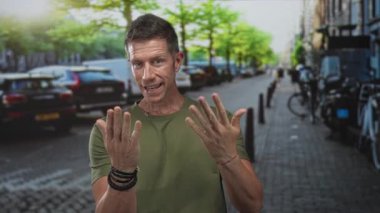 Adam, park etmiş arabalar ve bisikletlerle sokakta el kol hareketi yaparken, yeşil tişört ve bilezikler giyerken, dostça güven ve nişan gösterirken, avuç içi ve açık eller gösteriyor..