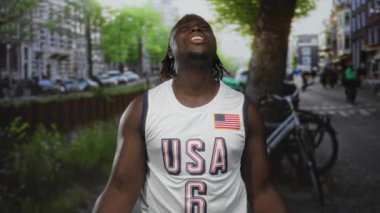 ABD basketbol formalı bir adam, yumruklarını sıkmış ve başını arkaya eğip bir caddeye bakıyor, zaferi kutluyor..