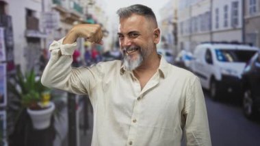 Adam kaslarını esnetiyor ve gülümsüyor, kollarını sıvayıp çıplak kollarını gösteriyor, dar bir şehir caddesinde park halindeki minibüslerle ve saksı bitkileriyle poz veriyor;.