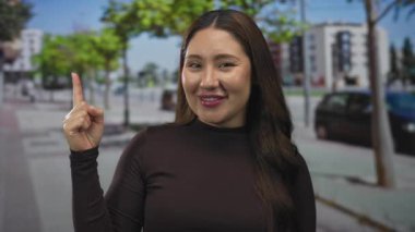 Latin kökenli genç bir kadın, park halindeki araba ve ağaçların yakınındaki şehir caddesinde işaret parmağını yukarı kaldırıyor..