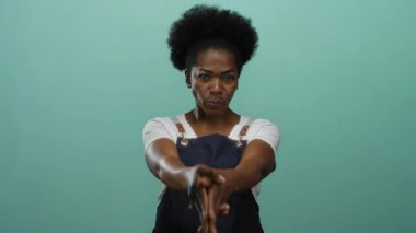 Önlüklü kadın, izole edilmiş yeşil arka plana parmak tabancası hareketi yapıyor. Basit bir stüdyo ortamında oyuncu ve kendinden emin bir ifade sergiliyor..