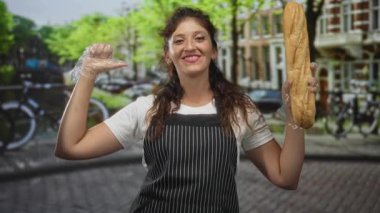 Çizgili önlüklü kadın, eldivenli el ve başparmağıyla bir baget tutuyor ve bisiklet ve tuğla evlerle dolu bir kanalın kenarındaki kaldırım taşı sokakta kendini işaret ediyor..