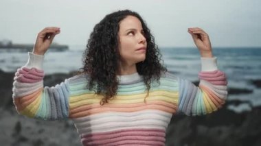 Kumsalda huzurlu bir anın tadını çıkaran kadın renkli süveter ve kıvırcık saçlarla, sakin bir okyanus zeminine karşı, deniz kenarında rahatlık ve özgürlük taşıyor..