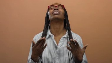 Gözlüklü kadın gömlek yakasını kahverengi izole bir arka plana göre ayarlıyor. Düşünceli bir ifade ve uzun kıvırcık saçla ısı rahatsızlığını gösteriyor..