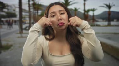 Kadın, palmiye ağaçlı bir caddede görünen yayaların olduğu krem rengi bir caddede yanaklarına yumruk atıyor; üzüntü..