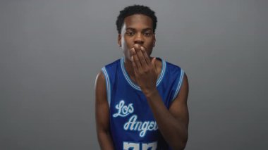 Mavi basketbol formalı Afrikalı Amerikalı bir adam. Gri bir stüdyo ortamında el pençe divan öpüşüyor..