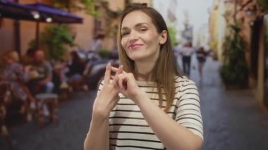 İşlek bir cadde terasında, şemsiyelerin altındaki kafe masalarının yanında neşeli bir gülümsemeyle işaret parmaklarını çapraz yapan bir kadın..