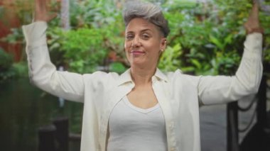 Kadın, yemyeşil bitkilerle dolu bir binanın avlusundaki çeşmenin yanında gülümseyerek kameraya doğru kollarını açar; neşe.