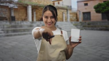 Önlüklü kadın garson bina yakınındaki taş basamaklarda not defteri ve kalem tutarken kamerayı işaret ediyor; dostça karşılama servisi.