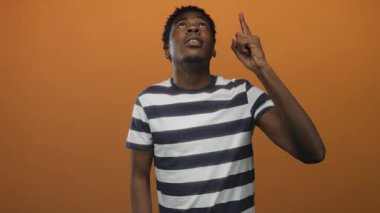 Young man in striped shirt pointing finger to object and gazing upward in orange studio; inspiration.