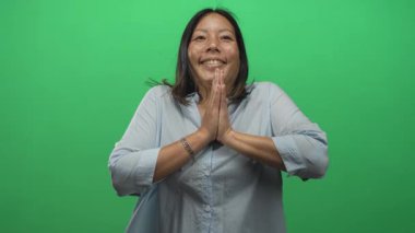 Yeşil bir stüdyoda dua jestinde elleri birbirine bastırılmış gülümseyen bir kadın; minnettarlık farkındalığı.