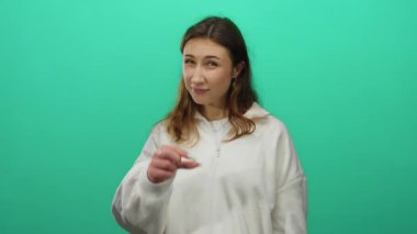 Parlak yeşil arka planda, parmakları küçük gösteren jestler yapan, beyaz bir kazak giyen ve neşeli, anlamlı bir poz veren bir kadın..