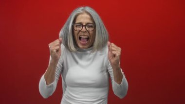 Yumruklarını sıkmış ve ağzı açık bir kadın stüdyoda kırmızı arka planda bağırıyor, gri saçlı, gözlüklü ve beyaz bluzlu;.