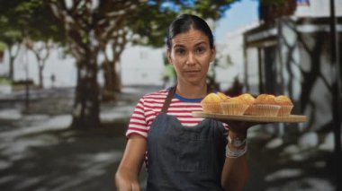 Kot önlüklü kadın elinde bir tepsi kek ve parmağıyla caddedeki kamerayı işaret ediyor..