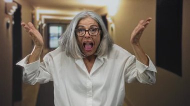 Bir binanın içindeki otel koridorunda el çırpan ve gülümseyen bir kadın, gri saçlı ve gözlüklü, beyaz gömlekli, kolları havada ve hareketli; neşe, sevinç, kutlama.